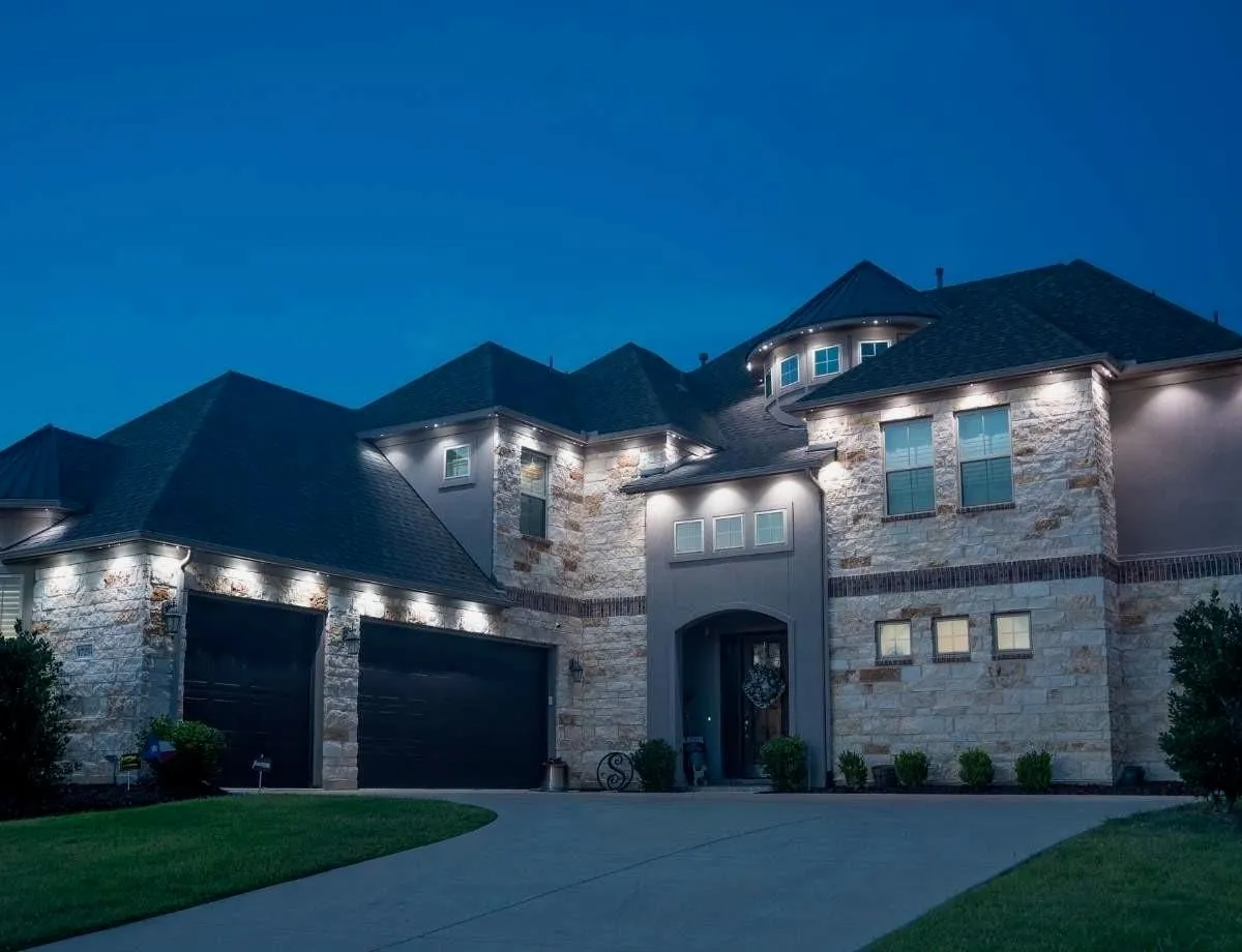 Large stone home at dusk with bright white lights washing down from the roofline and soffits.