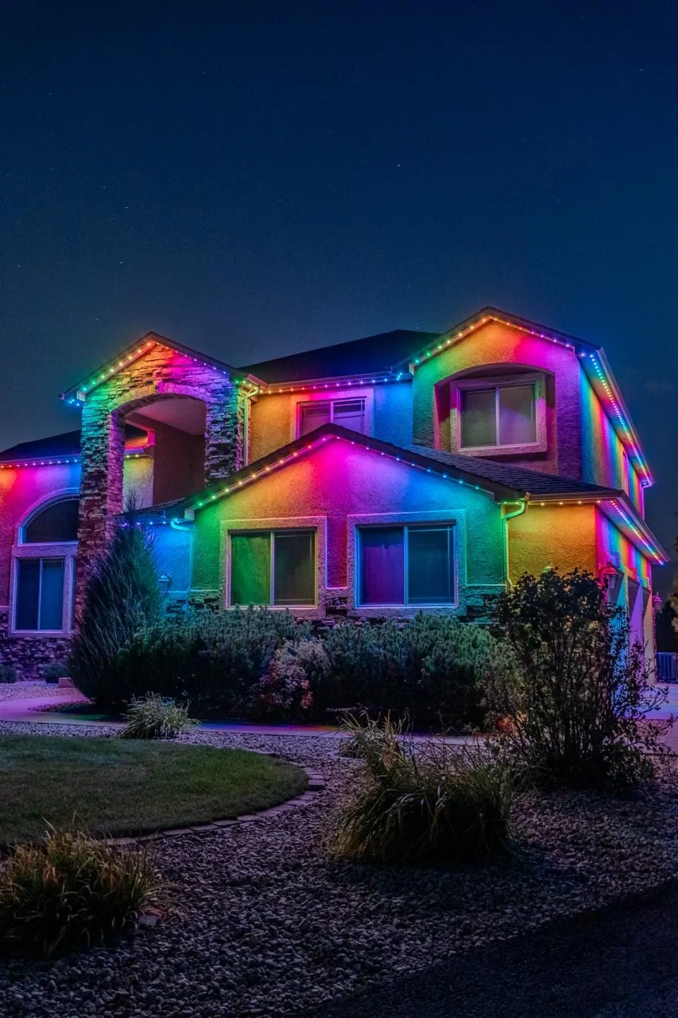 Two story stucco and stone home at night with a full color gradient of lights across every roofline.