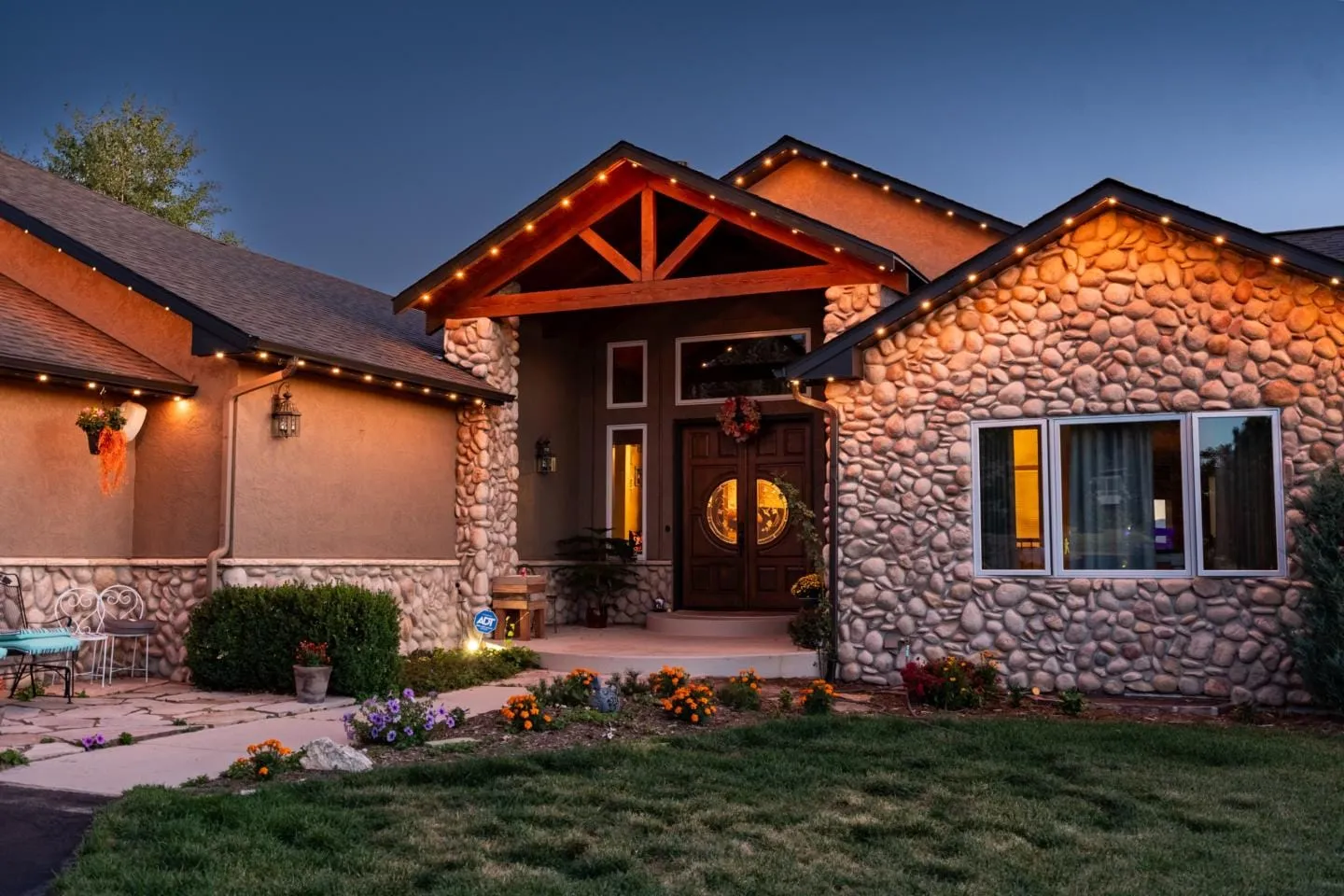 Stone and stucco home at dusk with warm white roofline lights tracing the gables and timber entry.
